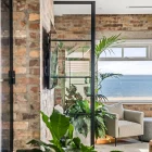 Bespoke steel bifold doors open to a light-filled space with a view of the sea, surrounded by greenery and textured brick walls.