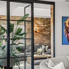 Steel-framed glass bifold doors open to a rustic living area with exposed brick walls, a plush sofa, and a large potted plant.