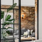 Black steel bifold doors open to a stylish living area featuring exposed brick walls, grey sofas, and a coffee table with flowers.