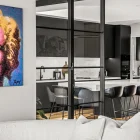 Black steel bifold doors separate a modern kitchen from a living area, featuring marble accents and sleek grey bar stools.