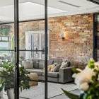 Bifold steel frame glass doors open into a modern living space with exposed brick, a grey sofa, indoor plants, and two dogs.