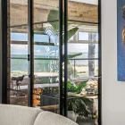 Black steel-framed bifold doors open to a spacious indoor plant area, revealing a scenic view outside. Textured walls and modern decor enhance the setting.