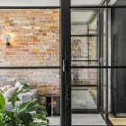 Interior view featuring steel frame glass bifold doors separating a modern living area with exposed brick walls and lush greenery.