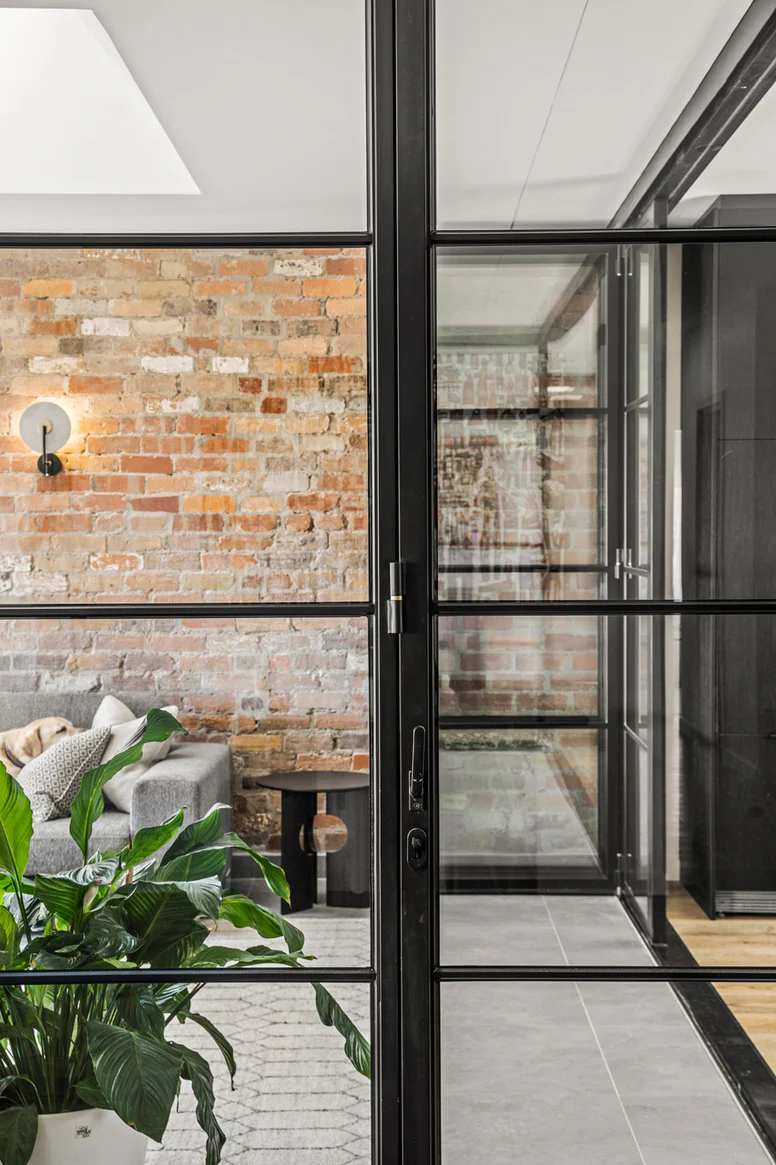 Interior view featuring steel frame glass bifold doors separating a modern living area with exposed brick walls and lush greenery.