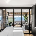 Black steel bifold doors open to a coastal view, showcasing a modern kitchen with marble countertops and sleek cabinetry, blending indoor and outdoor spaces.