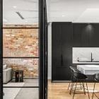 Steel frame bifold doors separate a modern kitchen with marble accents and black cabinetry from a stylish living area with exposed brick walls.