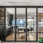 Black steel-framed bifold doors separate a modern dining area from a living space, featuring wood flooring and a touch of greenery.