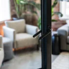 Black steel bifold door handle in focus, with a blurred background featuring plush grey armchairs and greenery, creating a modern interior vibe.