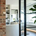 Black steel bifold doors open to a modern living space with a round white sofa, wooden flooring, and large windows, framed by brickwork.
