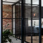 Black steel bifold doors with glass panels, framed against a rustic brick wall, reveal a modern kitchen space and indoor plant.