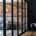 Black steel bifold doors separate a modern room with exposed brick walls and a sleek grey sofa, enhancing the architectural design.