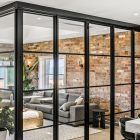 Black steel-framed glass bifold doors open to a modern living room with a grey sofa, exposed brick walls, and natural light.