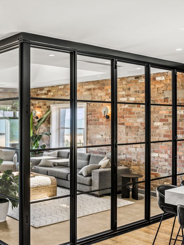 Black steel-framed glass bifold doors open to a modern living room with a grey sofa, exposed brick walls, and natural light.