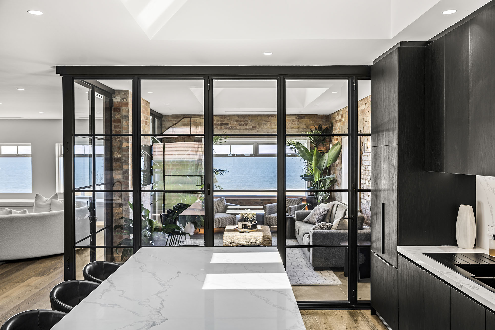 Steel frame glass bifold doors open to a bright, stylish living space with a view of the water, featuring neutral tones and modern decor.