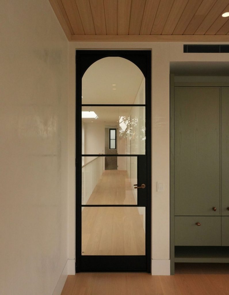 Black steel-framed door with arched top, leading to a light-filled corridor; soft neutral walls and wooden ceiling enhance the modern design.