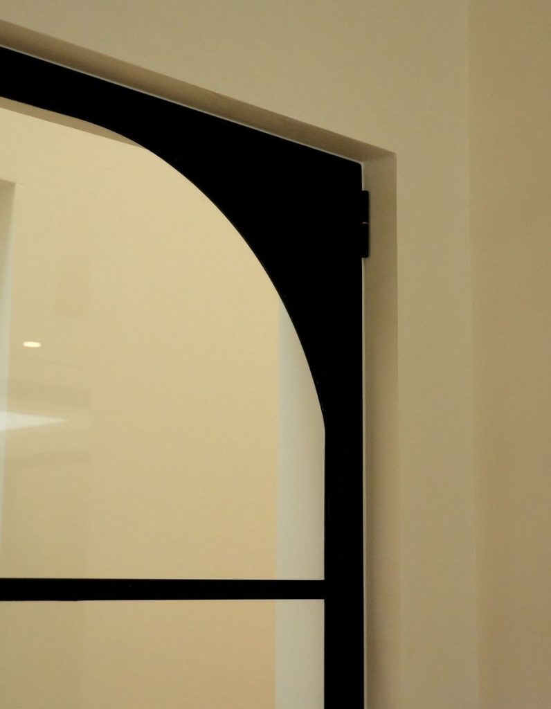 Bespoke black steel door with an arched top, set within a light-coloured wall, highlighting minimalist design and craftsmanship.