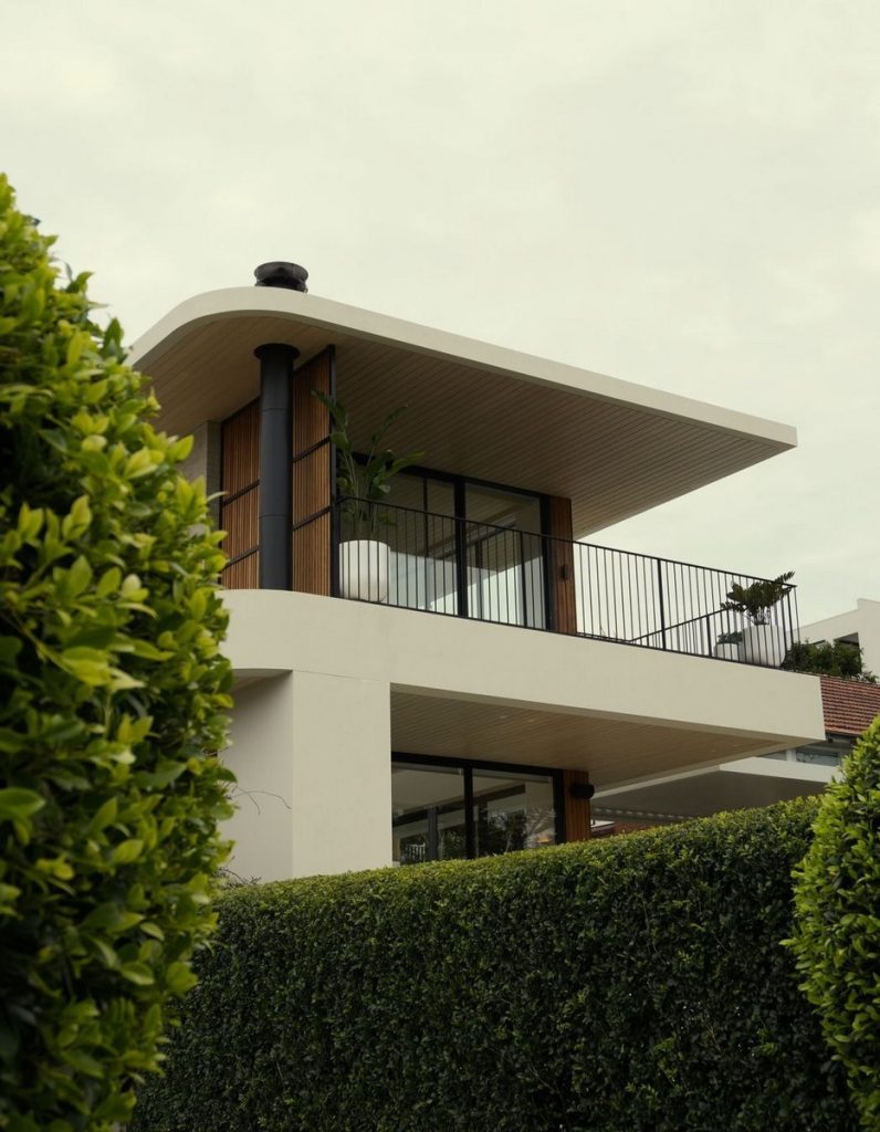 Modern two-storey house with a curved balcony, featuring large glass windows, warm timber accents, and neatly trimmed greenery.