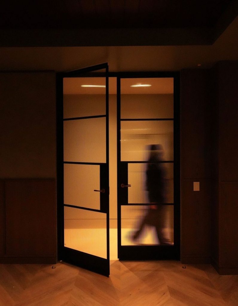 Bespoke steel doors with intricate glass panels, framed in black, creating a warm, softly lit interior with a figure silhouetted in motion.