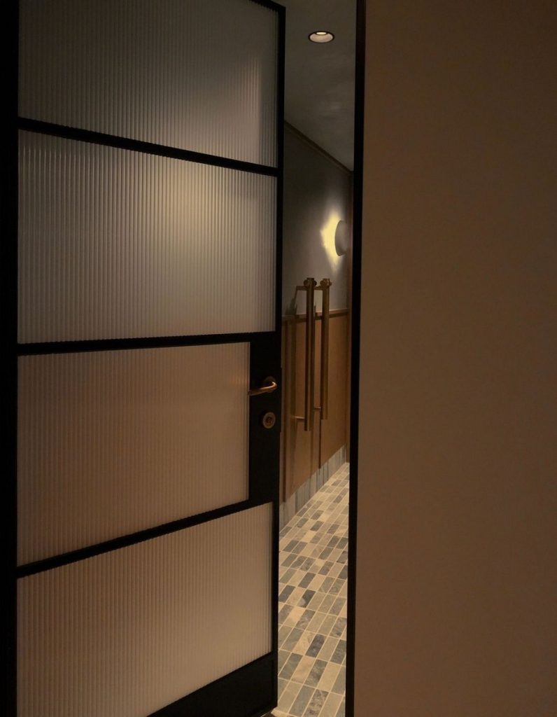 Interior view of a sleek black-framed door with frosted glass panels, leading into a warmly lit corridor with tiled flooring.