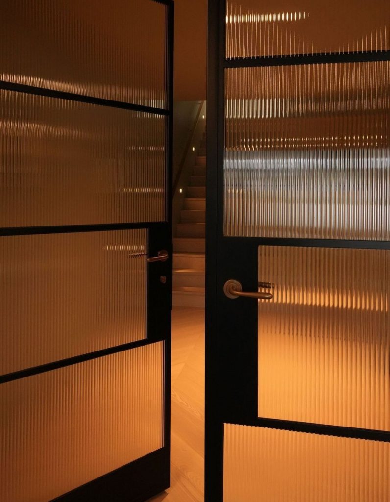 Framed glass doors with ribbed panels open to reveal a softly lit staircase, exuding warmth and modern elegance.