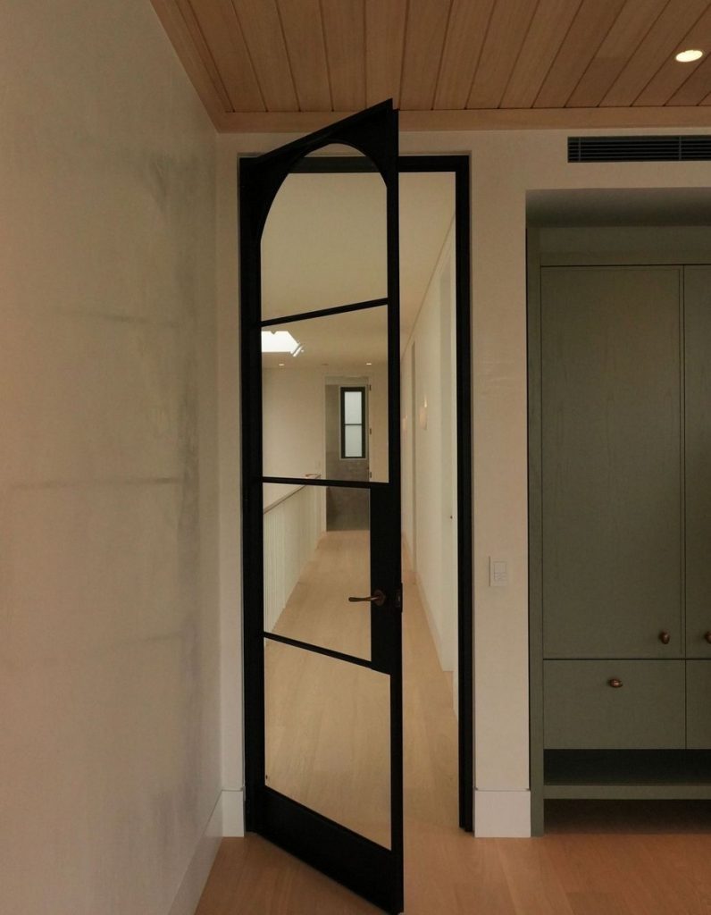 Black arched steel door open to reveal a light-filled hallway, featuring muted tones and a wooden ceiling, enhancing modern elegance.