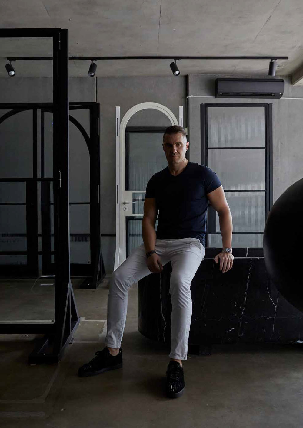 A man in a navy t-shirt and white trousers sits on a dark stone surface, surrounded by elegant steel door designs in a modern setting.