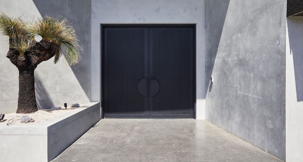 Bespoke black steel double doors set against a textured concrete wall, framed by a minimalist landscape with a palm tree and sandy area.