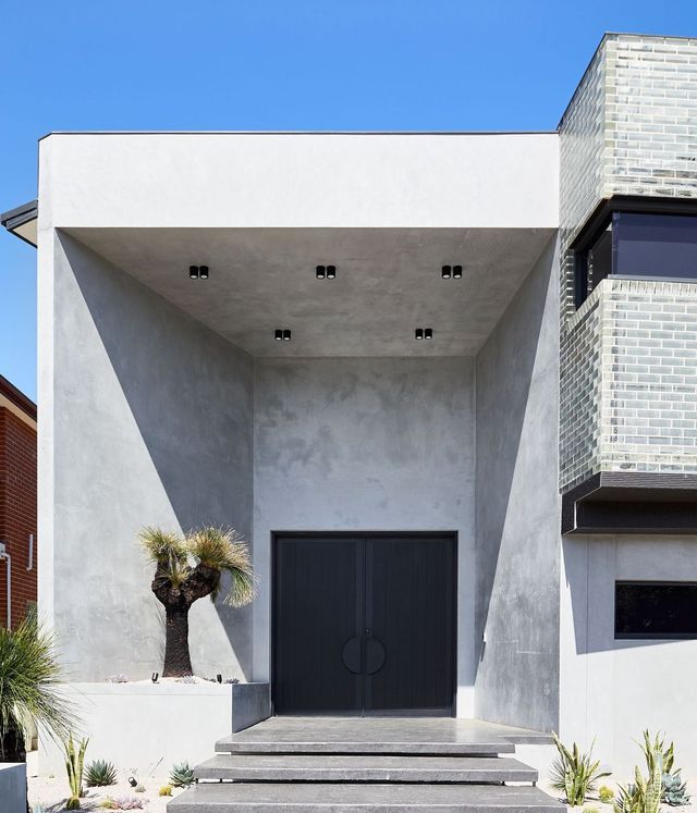 Benfield Concrete House Entrance with Steel Doors and Modern Design Modern concrete house entrance featuring large dark double doors, a palm tree, and angular architectural lines against a clear blue sky.