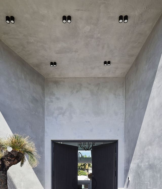 Benfield Concrete House Entrance with Steel Door and Modern Design Open entrance of a concrete house featuring minimalist design, grey walls, modern ceiling lights, and a glimpse of greenery outside.