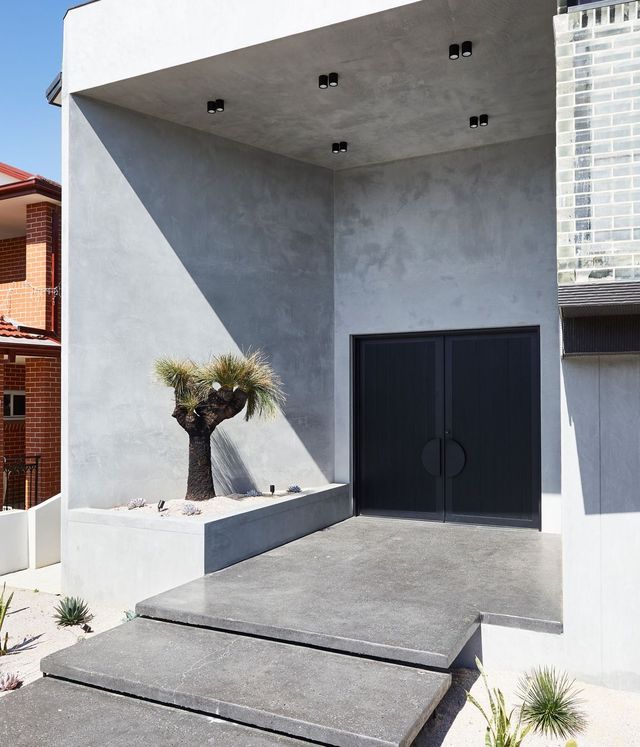 Benfield Concrete House Entrance with Steel Doors and Sleek Design A modern concrete entrance features a dark double door, a textured wall, and a neatly landscaped pathway with a palm tree.