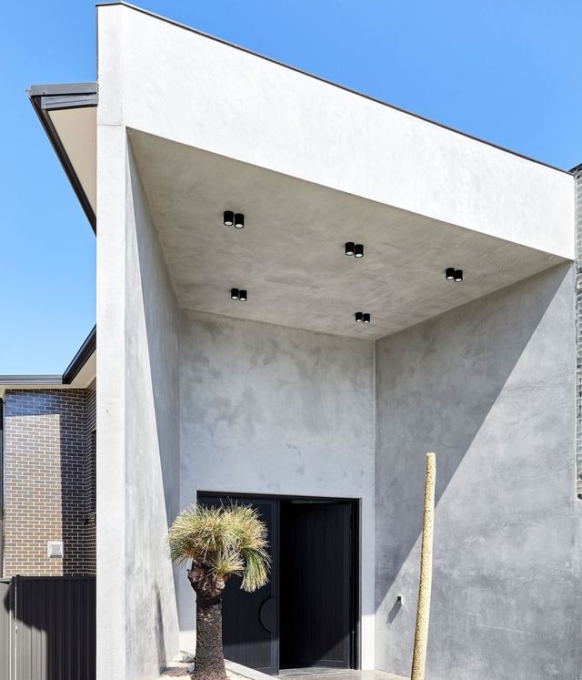 Benfield Concrete House with Bespoke Steel Door Design Modern concrete house entrance with a striking angular roof, featuring recessed lighting and a tall palm tree beside the doorway.