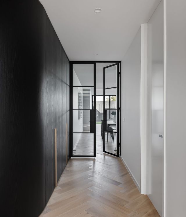 Modern hallway featuring a black wall and bespoke black steel-framed glass doors, with herringbone wooden flooring and contemporary design.