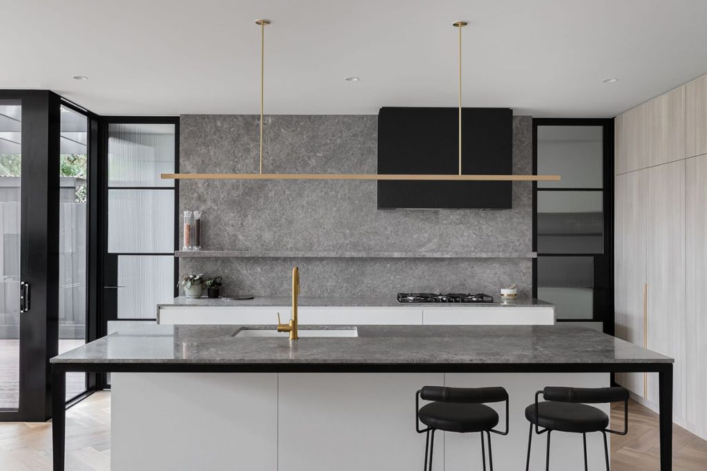 Modern kitchen featuring a large grey stone island with a sink, minimalist cabinetry, sleek black accents, and pendant lighting.
