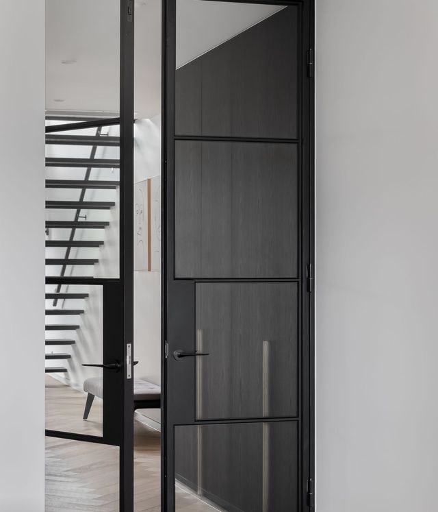 Sleek black steel-framed door with glass panels, leading into a contemporary interior featuring wooden flooring and a modern staircase.