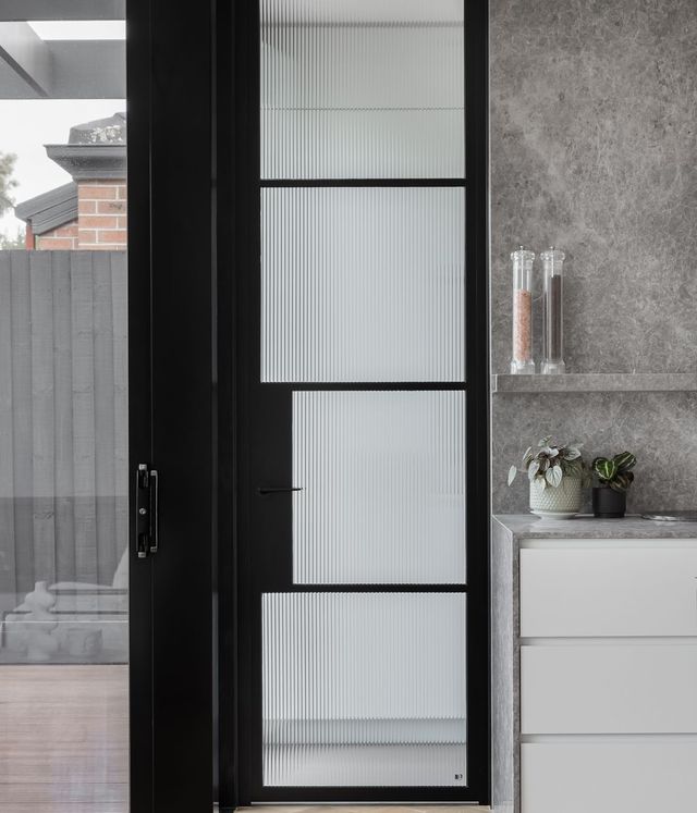 Bespoke black steel-framed door with ribbed glass panels, set against a grey stone wall and minimalist kitchen with potted plants.