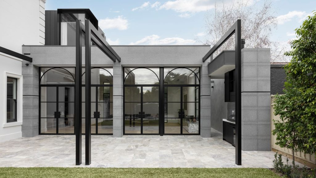 Modern architectural design featuring a grey stone building with large arched black-framed glass doors, surrounded by greenery and paving.