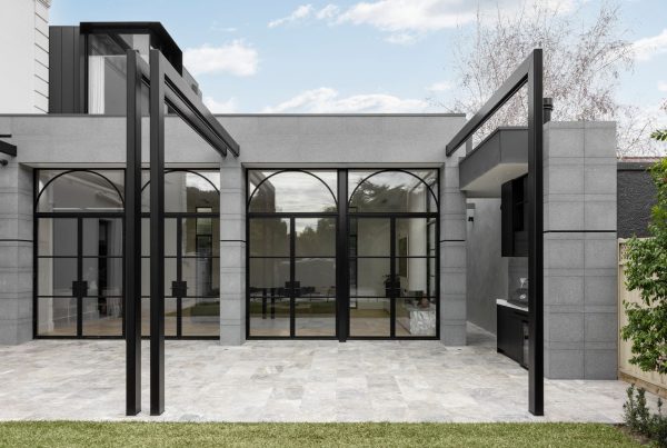 Modern architectural design featuring a grey stone building with large arched black-framed glass doors, surrounded by greenery and paving.