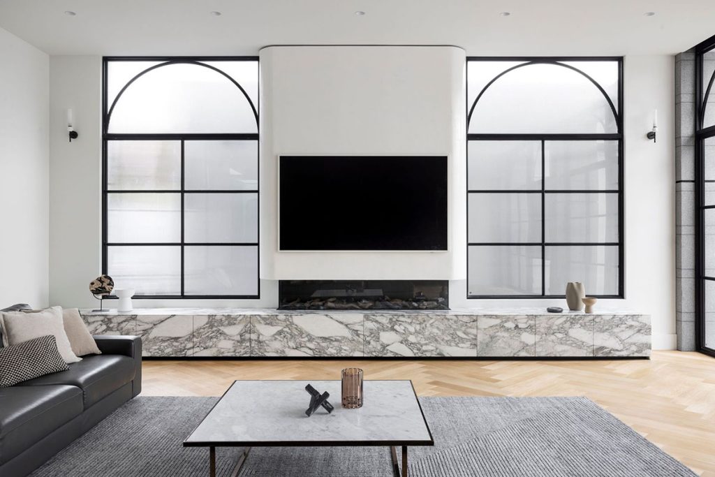 Modern living room featuring a sleek marble media unit beneath a large television, framed by elegant arched steel windows and a minimalist grey sofa.