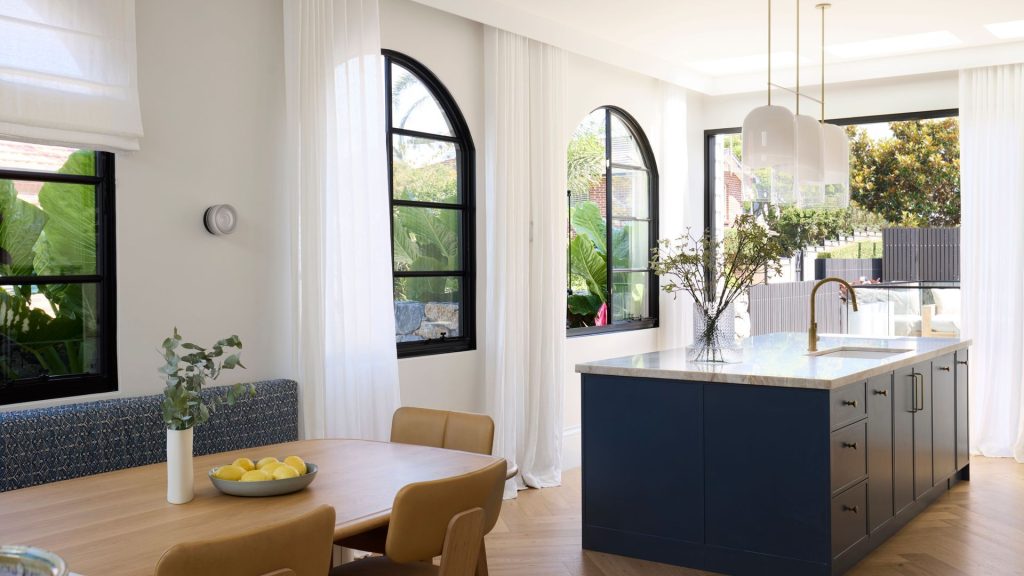 Bright modern kitchen featuring elegant steel-framed windows, a large island with a marble top, light wooden dining furniture, and greenery.