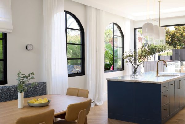 Bright modern kitchen featuring elegant steel-framed windows, a large island with a marble top, light wooden dining furniture, and greenery.