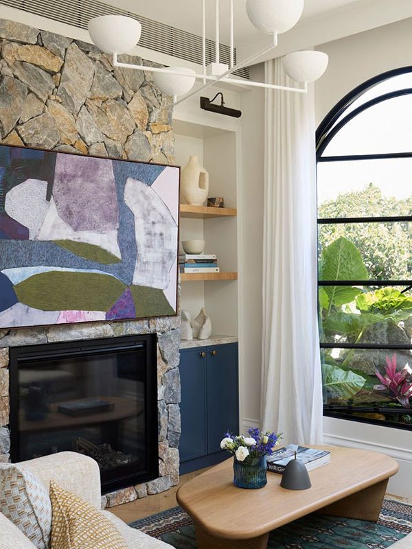 Modern living room featuring a stone fireplace, large arched window with steel framing, abstract artwork, and neutral furnishings.