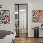 Steel frame glass sliding doors separate a modern dining area from a hallway, featuring wooden flooring and artworks, with a dog resting nearby.