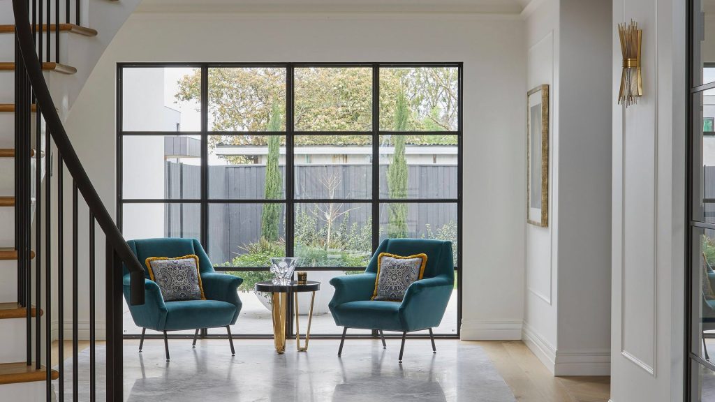 Bright foyer featuring two teal armchairs with decorative cushions, a small side table, and large steel-framed glass doors leading to a garden.