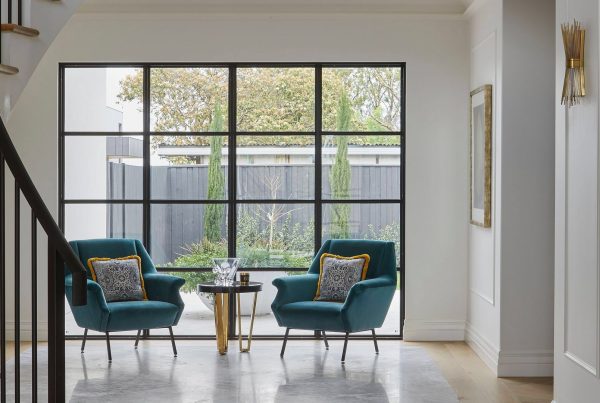Bright foyer featuring two teal armchairs with decorative cushions, a small side table, and large steel-framed glass doors leading to a garden.