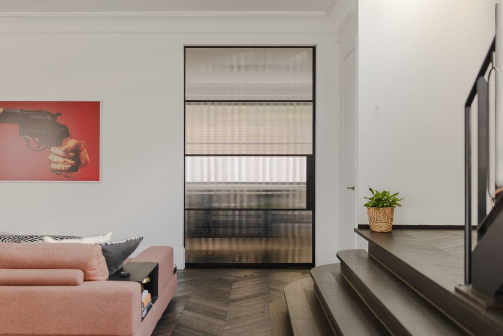 Contemporary interior featuring a steel-framed glass door, a stylish sofa, a bold artwork, and a staircase with dark wood flooring.