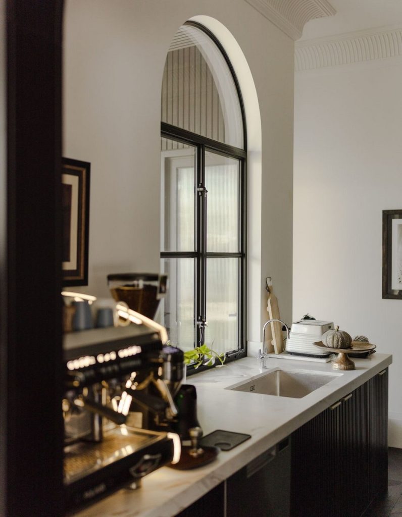 A stylish kitchen featuring a steel-framed arched window, dark cabinetry, a marble countertop, and a coffee machine inviting a modern aesthetic.