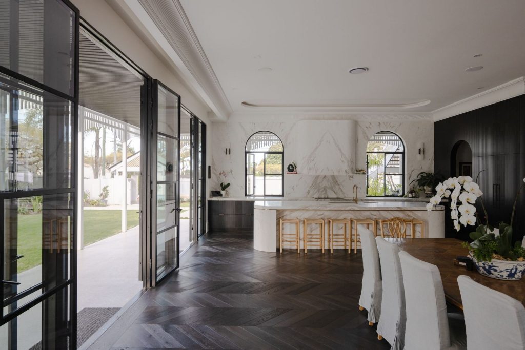 Bright open-plan interior featuring a marble kitchen with black steel-framed doors, stylish dining area, and large windows overlooking the garden.