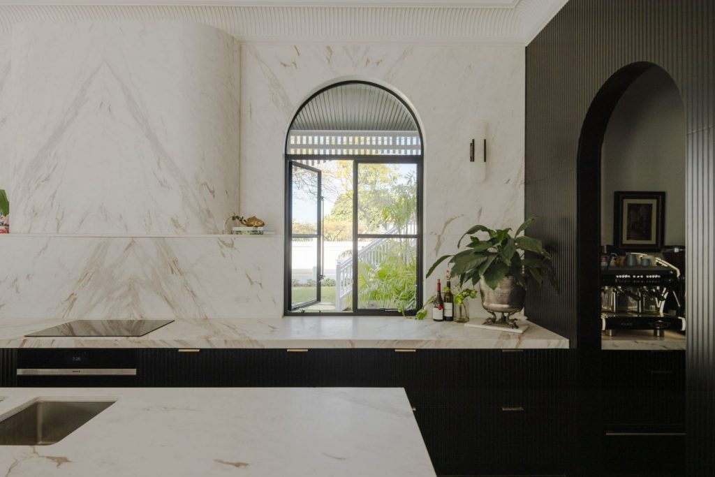 Elegant kitchen with marble surfaces, featuring a large arched window framed in steel, overlooking a lush outdoor view and greenery.