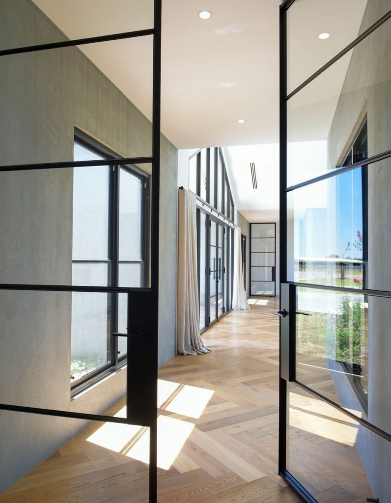 Elegant interior featuring black steel-framed doors and windows, minimalist design, warm wood herringbone flooring, and soft curtain drapes.