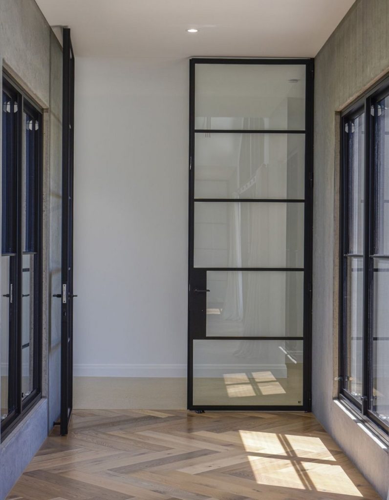 Contemporary corridor featuring steel-framed doors and windows, grey walls, and herringbone wooden flooring, creating an elegant interior.